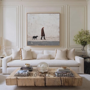 Könnte beinhalten: Ein quadratisches gerahmtes Wandbild mit einem Gemälde einer Person in einem braunen Mantel, die einen schwarzen Hund an einer hellen Wand entlang führt. Das Kunstwerk hängt über einem weißen Sofa mit beigen Kissen und einem massiven Holztisch.