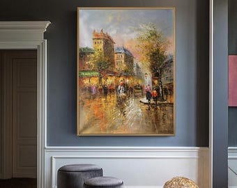 May include: An oil painting in a gold frame depicting a bustling city street scene with tall buildings, trees, and figures walking along a wet pavement. The artwork features warm yellow, orange, and brown tones with visible brushstrokes.