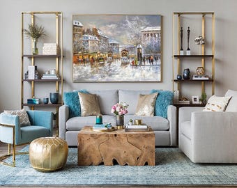 May include: A living room featuring a grey sofa with teal and gold cushions, a natural wood block coffee table, two gold metal shelving units, a teal armchair, a gold pouf, and a large framed oil painting of a snowy city street scene.