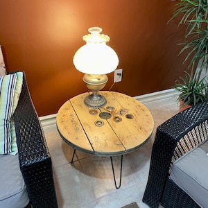 Peut inclure: Une table en bois en forme de bobine avec des pieds en épingle à cheveux noirs et une lampe blanche avec un abat-jour en verre. La table est posée sur un sol carrelé avec un tapis devant elle.