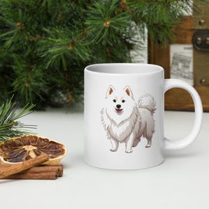 Może przedstawiać: Biały ceramiczny kubek z kreskówkową ilustracją puszystego, białego psa rasy samojed. Pies ma uśmiechniętą minę i stoi wyprostowany. Kubek ma standardowy uchwyt i świąteczne tło.
