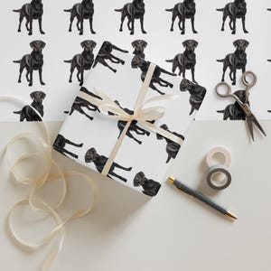 May include: White wrapping paper featuring a repeating pattern of black Labrador Retrievers. The wrapped gift is tied with a cream-colored ribbon. Scissors, tape, and a pen are also visible.