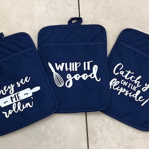 May include: Three navy blue quilted oven mitts with white text. One says "They see me rollin'" with a rolling pin graphic. Another says "Whip it good" with a whisk. The third says "Catch you on the flipside" with a spatula.
