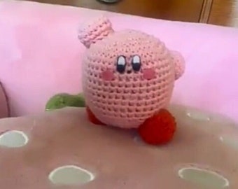 May include: A handmade pink crochet Kirby plushie with black eyes, pink cheeks, and red feet. The plushie is sitting on a light pink surface. The plushie is a popular character from the video game series.