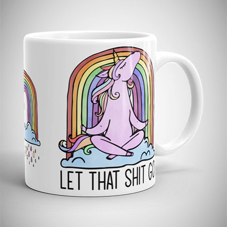 May include: White ceramic mug featuring a cartoon unicorn meditating in front of a rainbow. The unicorn is pink with a yellow horn and colorful mane. The text "LET THAT SHIT GO" is printed below the unicorn.