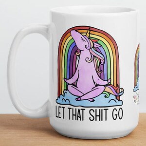May include: White ceramic mug with a whimsical design. The mug features a meditating pink unicorn under a rainbow, with the text "LET THAT SHIT GO" in black. The mug has a large handle and is suitable for hot or cold beverages.