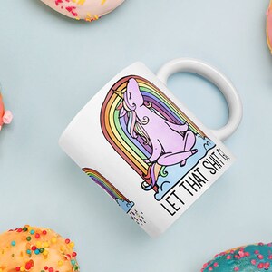 May include: White ceramic mug featuring a cartoon unicorn sitting on a rainbow with the text "LET THAT SHIT GO". The unicorn is pink with a yellow horn and colorful mane. The mug is surrounded by donuts.