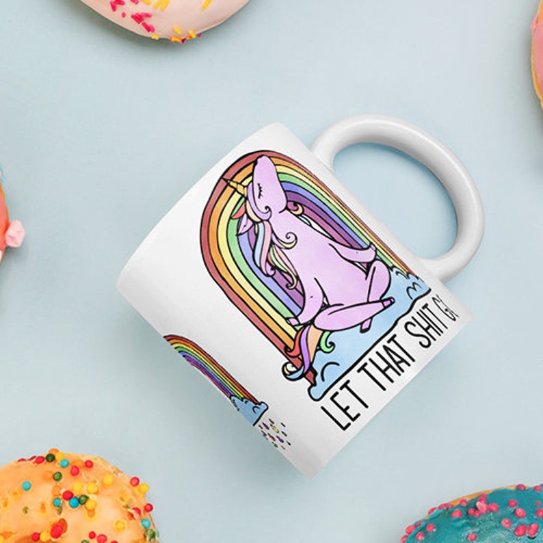 May include: White ceramic mug featuring a cartoon unicorn sitting on a rainbow with the text "LET THAT SHIT GO". The unicorn is pink with a yellow horn and colorful mane. The mug is surrounded by donuts.