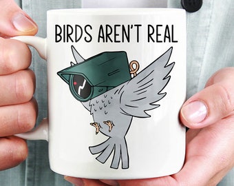 Könnte beinhalten: Weiße Keramik-Tasse mit dem Text "BIRDS AREN'T REAL" und einer Cartoon-Illustration eines grauen Vogels mit einer grünen Überwachungskamera. Die Tasse hat einen weißen Henkel.