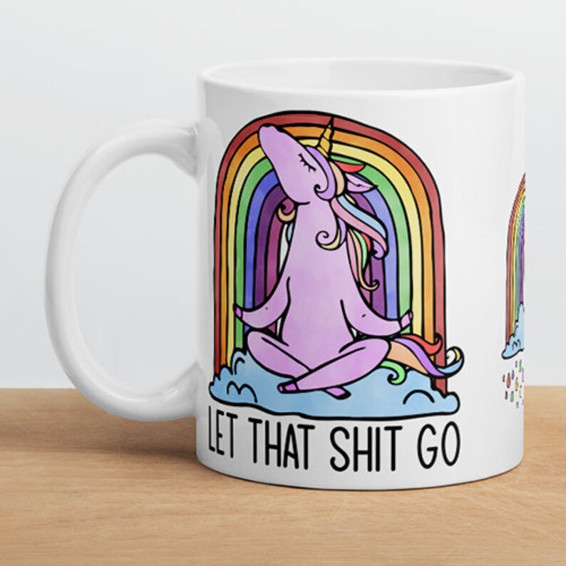 May include: White ceramic mug featuring a cartoon unicorn meditating in front of a rainbow. The unicorn is purple with a golden horn and colorful mane and tail. The text "LET THAT SHIT GO" is printed below the unicorn.