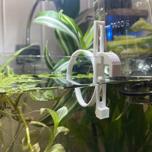 May include: A white 3D-printed aquarium filter holder, partially submerged in water. The holder has a circular design with a vertical support structure. Green aquatic plants and floating plants are visible in the water, with a glass tank in the background.
