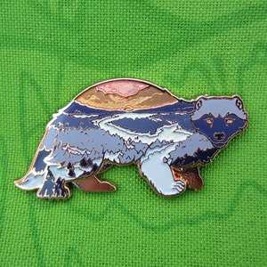 Puede incluir: Un pin de esmalte detallado con forma de tejón, con un diseño de paisaje. El pin muestra una puesta de sol sobre montañas y un río, con tonos azules, grises y cobrizos. El pelaje del tejón está representado en varios tonos de gris y blanco.