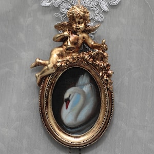 Könnte beinhalten: Eine goldfarbene Cherubfigur mit Trompete über einem ovalen Rahmen. Der Rahmen enthält ein Gemälde eines weißen Schwans mit rotem Schnabel. Der Rahmen ist mit Rosen verziert. Aufgehängt an einem Spitzendetail.