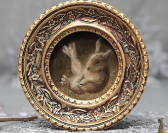 May include: A circular, ornate gold-toned frame featuring intricate floral and leaf relief carvings. The center contains a small, detailed painting of a brown rabbit curled in a resting position against a dark, muted green background.