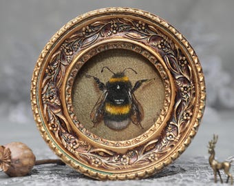Puede incluir: Un marco redondo y ornamentado de tono dorado con detalles florales en relieve, que encierra una representación bordada detallada de un abejorro con rayas negras y amarillas, alas translúcidas y antenas, sobre un fondo verde oliva apagado.