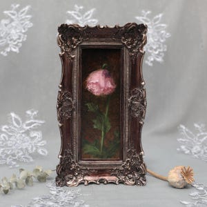 Può includere: Un dipinto rettangolare di un singolo papavero rosa con foglie e stelo verdi, su uno sfondo marrone scuro. L'opera è racchiusa in una cornice ornata color bronzo scuro con dettagli in rilievo raffiguranti motivi floreali e volute.