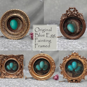Puede incluir: Pinturas originales de huevos azules enmarcadas. La obra presenta un huevo o huevos turquesa vibrantes anidados en un nido de plumas, enmarcados en marcos dorados o bronce ornamentados. El texto "Original Blue Egg Painting Framed" es visible.