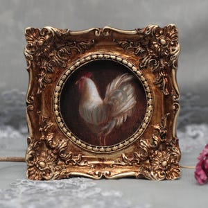 Può includere: Dipinto incorniciato di un gallo bianco con cresta rossa, su sfondo scuro. L'opera d'arte è racchiusa in una cornice quadrata dorata e ornata con dettagliati disegni floreali e a volute, creando un'estetica vintage.