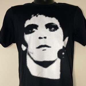 以下が含まれることがあります： 白黒のピクセル化された人物の顔の画像がプリントされた黒いTシャツ