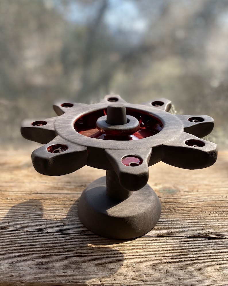 May include: A black ceramic candle holder with eight candle slots and a central, raised, circular platform. The candle holder has a starburst design and a red glaze on the inner rim of the candle slots.