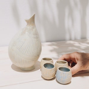 Può includere: Un set di quattro piccole tazze in ceramica bianca con una glassa blu chiaro e un design testurizzato, accanto a un'anfora in ceramica bianca più grande con una glassa e un design simili.
