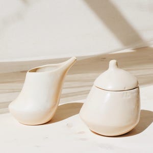 Puede incluir: Un juego de jarra de crema y azucarero de cerámica blanca. La jarra de crema tiene un pico curvo y un cuerpo redondeado. El azucarero tiene una tapa y un cuerpo redondeado.