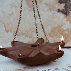 May include: A terracotta oil lamp with five wicks hangs from a chain. The lamp is lit and the flames are visible. The lamp is hanging against a light brown background.