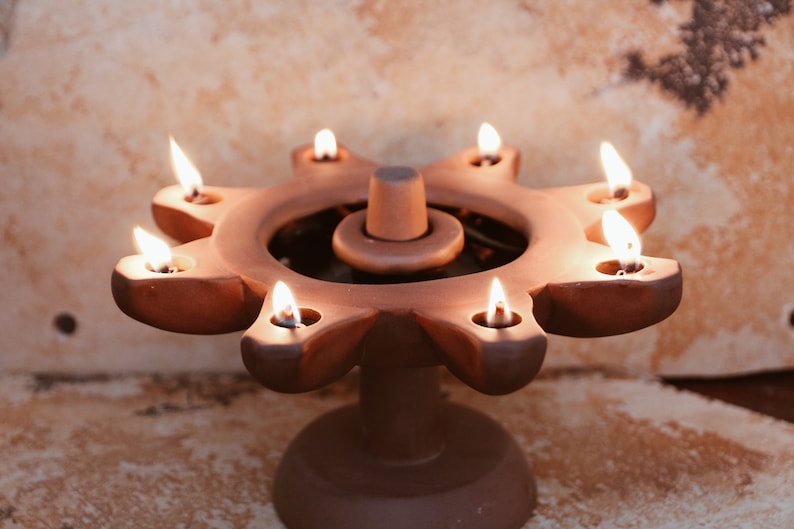 May include: A terracotta candle holder with seven open flames, shaped like a sun with a central circular opening. The holder is on a round base.