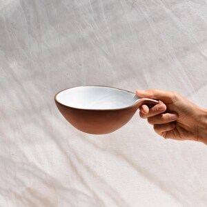 May include: A white and brown ceramic bowl with a unique handle. The bowl is being held by a hand.