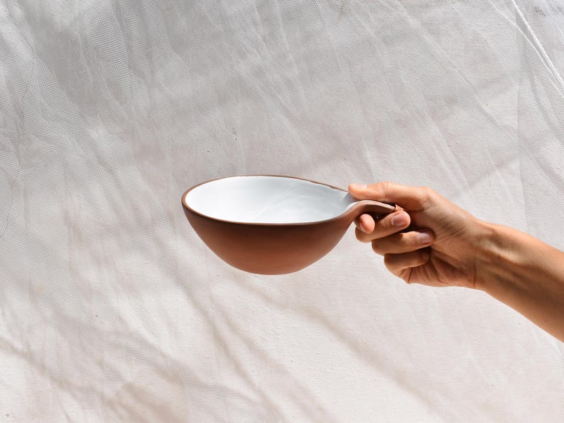 May include: A white and brown ceramic bowl with a unique handle. The bowl is being held by a hand.