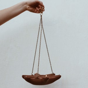 May include: A terracotta hanging oil lamp with a star-shaped design and three chains. The lamp is suspended from a hook.