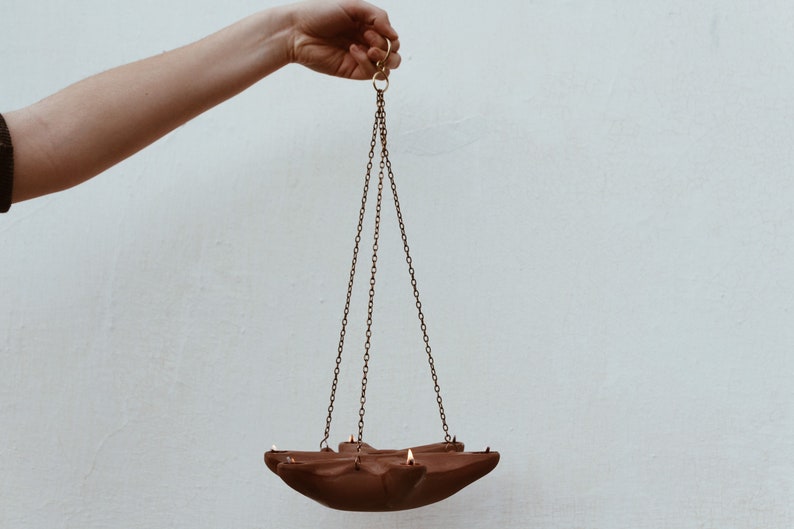 May include: A terracotta hanging oil lamp with a star-shaped design and three chains. The lamp is suspended from a hook.