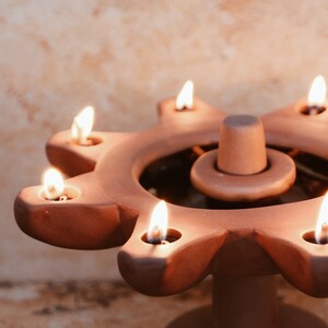 May include: A terracotta candle holder with seven candle slots, each with a lit candle. The holder is shaped like a star with a central circular base.