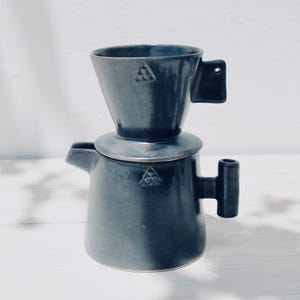 Puede incluir: Juego de café pour-over de cerámica azul oscuro. El juego incluye un vertedor en forma de cono, una tapa y una jarra con asa. Cada pieza tiene un diseño triangular. El juego está sobre una superficie blanca.