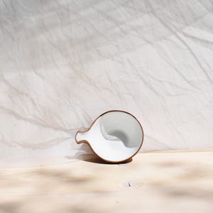 May include: A white ceramic pitcher with a brown rim, resting on a light wood surface. The pitcher is tilted slightly, revealing the inside.