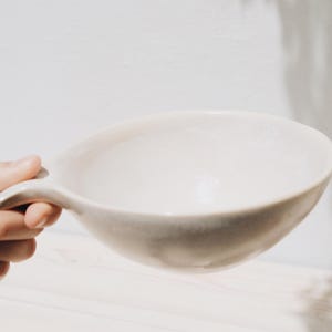 May include: A white ceramic bowl with a handle. The bowl is empty and has a slightly textured surface.