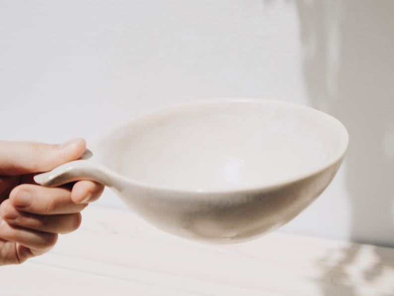 May include: A white ceramic bowl with a handle. The bowl is empty and has a slightly textured surface.