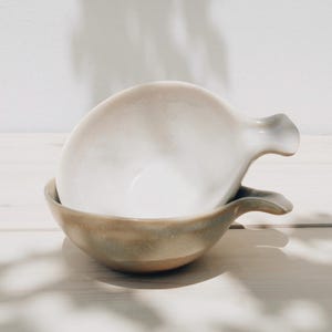 May include: Two ceramic bowls with a light brown glaze and a white interior. The bowls have a unique shape with a handle on one side.