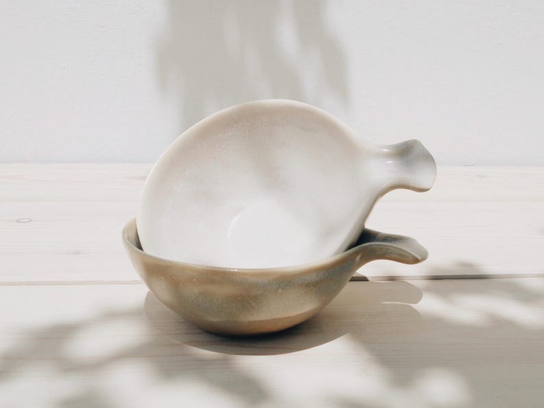 May include: Two ceramic bowls with a light brown glaze and a white interior. The bowls have a unique shape with a handle on one side.