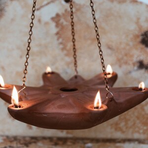 May include: A terracotta oil lamp with five wicks, hanging from a chain. The lamp is lit and the flames are casting a warm glow.