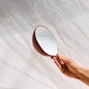 May include: A white ceramic bowl with a brown rim and a unique curved handle. The bowl is held in a hand against a white background.