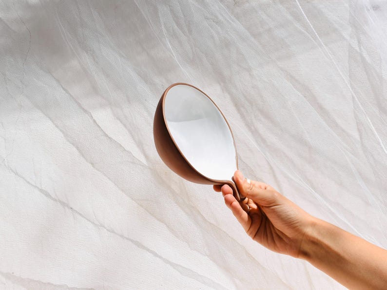 May include: A white ceramic bowl with a brown rim and a unique curved handle. The bowl is held in a hand against a white background.