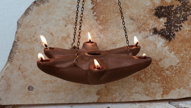 May include: A terracotta oil lamp with five wicks hanging from a chain. The lamp is lit and the flames are visible.
