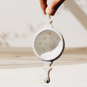 May include: White ceramic tea infuser with a handle and a round, perforated bottom. The infuser is held by a hand.