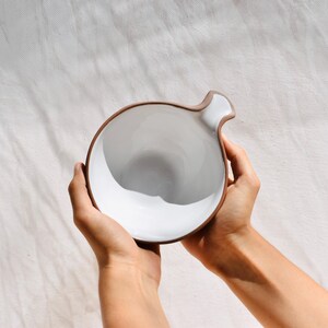 May include: A white ceramic bowl with a brown rim and a unique handle. The bowl is held in two hands.