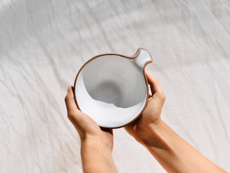 May include: A white ceramic bowl with a brown rim and a unique handle. The bowl is held in two hands.