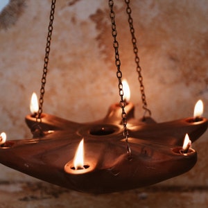 May include: A terracotta oil lamp with six wicks hangs from a chain. The lamp is lit and the flames are casting a warm glow.