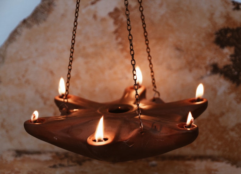 May include: A terracotta oil lamp with six wicks hangs from a chain. The lamp is lit and the flames are casting a warm glow.