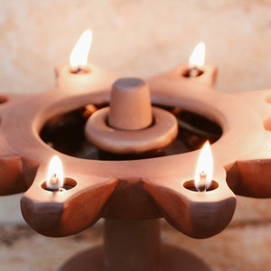 May include: A terracotta candle holder with seven lit wicks, shaped like a sun with a central circular well. The holder is on a textured surface.