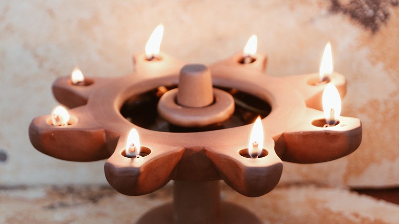 May include: A terracotta candle holder with seven lit wicks, shaped like a sun with a central circular well. The holder is on a textured surface.